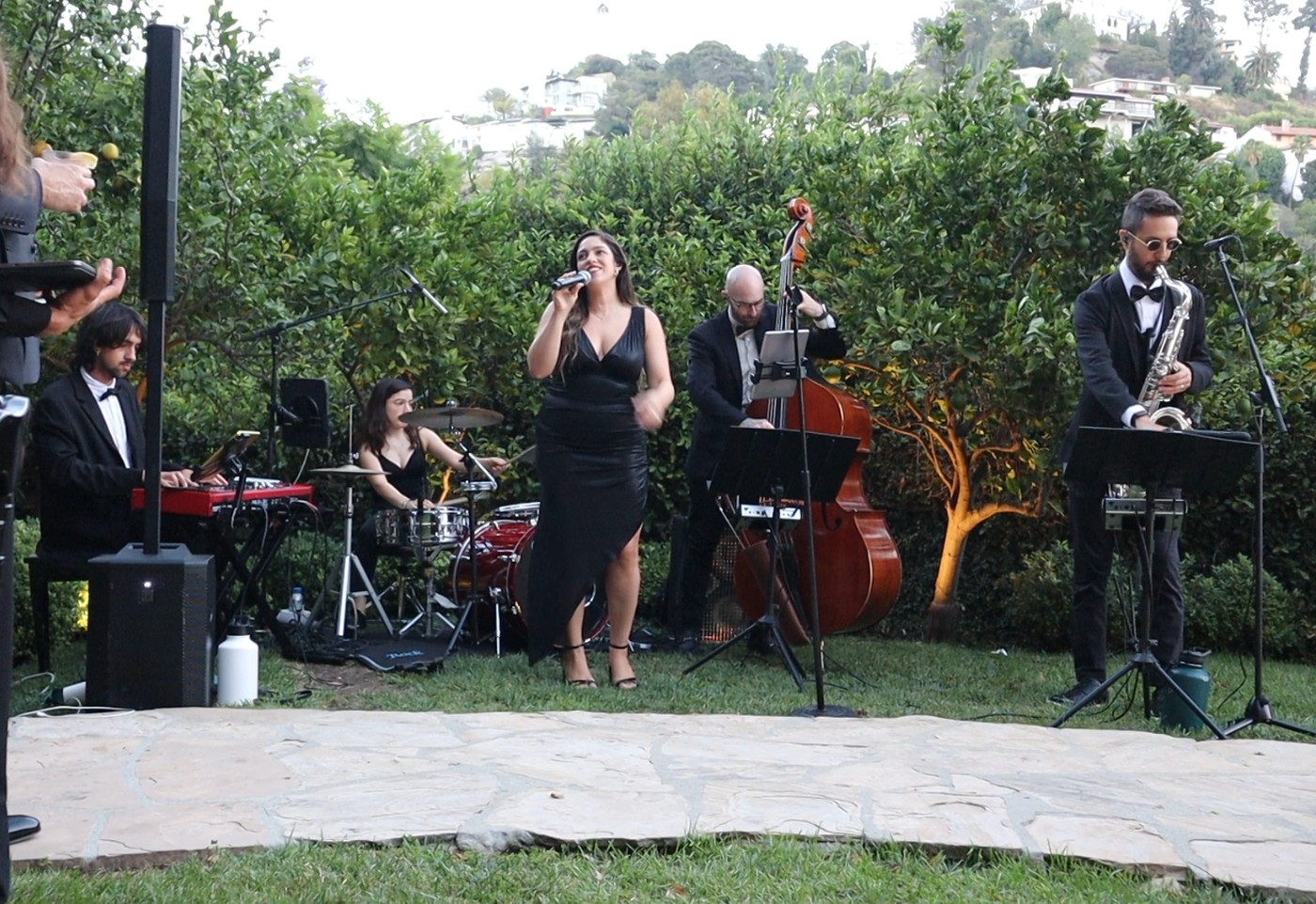 Live band with vocalist, saxophone, upright bass, keys and drums performing on a garden lawn at a hillside Los Angeles wedding venue, with speakers on stands