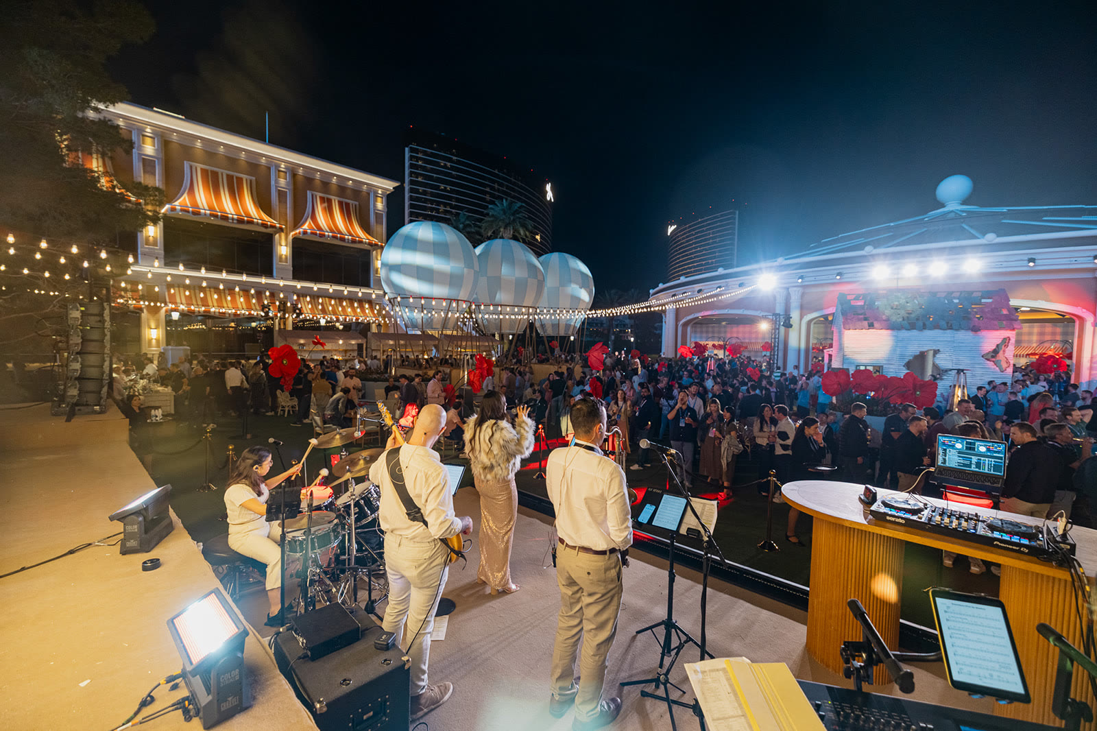Live band on an outdoor stage playing to a large corporate event crowd at night, with a DJ booth beside the stage